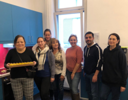 sechs Frauen und ein Mann stehen in der Küche, eine von den Frauen hält ein Tablett mit Kuchen in der Hand