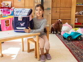 Ein kleines Mädchen sitzt an einem Tisch, auf dem ihr Schulranzen steht. Das Zimmer ist mit Spielsachen angefüllt