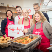 Gruppenbild Haya Molcho beim Samariter Suppentopf, mehrere Personen, tragen alle rote Samariter Suppentopf Schürzen, halten Schild in Hand, davor Essen in Wärmebehältern