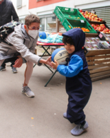 Bezirksvorsteherin 15. Bezirk in Sozialmarkt Pillergasse, kniet Richtung Boden und gibt Kind in Jacke mit Kapuze über dem Kopf Ball, im Hintergrund Kisten mit Obst