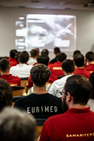 Aufnahme während einer Fortbildung, Teilnehmer:innen von hinten zu sehen, tragen T-Shirts mit Aufdruck European Medics und Samariterbund Wien, blicken auf Leinwand auf der Präsentation