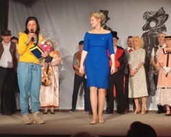 Eine Dame in einem blauen Kleid steht auf der Bühne, eine Dame mit gelbem T-Shirt spricht in ein Mikrofon.