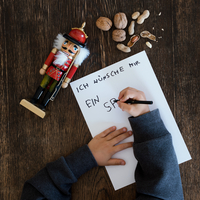 Aufnahme von oben, Kinderhand die einen Wunschzettel schreibt, mit Stift auf Papier, daneben weihnachtlicher Nussknacker und Walnüsse