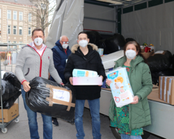 vier Personen mit FFP2-Masken mit Spielzeug in der Hand, dahinter LKW mit weiteren Sachspenden