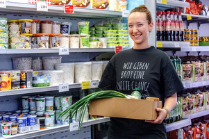 Mitarbeiterin aus Sozialmarkt mit blauem T-Shirt mit Aufdruck Wir geben Brot eine zweite Chance, steht in Sozialmarkt, hält Karton mit Lebensmitteln und Gemüse in der Hand, dahinter Kühlschrank mit weiteren Produkten und Regal mit Produkten, Haare zu einem Dutt zusammengebunden, lächelt in Kamera