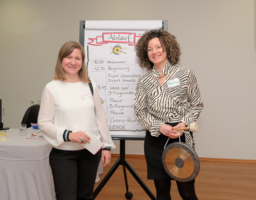 Zwei Frauen vor Flipchart, lächeln, eine hält Gong in der Hand