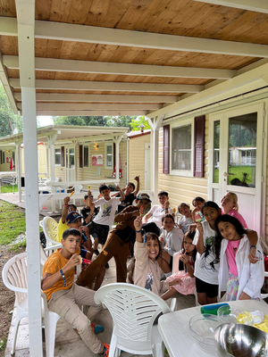 Gruppenbild viele Kinder auf einer überdachten Terrasse eines Holzhauses, Feriencamp, sitzen auf Plastiksessel oder stehen, halten Eis in die Hand und blicken lachend in Kamera 
