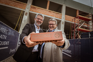 Oliver Löhlein und Stadrat Peter Hacker vor einer Baustelle, halten einen Ziegelstein gemeinsam in der HAnd und richten diesen in die Kamera, beide blicken in Kamera und lächeln