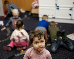 Innenaufnahme aus Ukraine Flüchtlingseinrichtung, im Vordergrund kleines Mädchen im rosa Shirt, dass direkt in die Kamera blickt, im Hintergrund weitere Kinder, die meisten von hinten fotografiert, die am Boden spielen