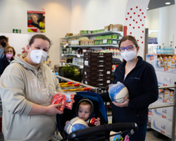 Mutter und Leiterin Sozialmarkt in Sozialmarkt, beide tragen FFP2-Maske und halten Bälle und andere Sportatikel in der Hand, in der Mitte Kleinkind im Kinderwagen, hält auch Ball in der Hand, im Hintergrund Supermarktatmosphäre mit Regalen voll Lebensmittel