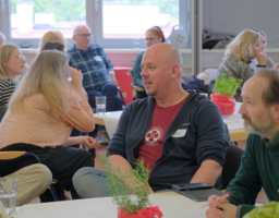 Gruppe von Menschen an Tischen, zwei Personen von vorne, Diskussion