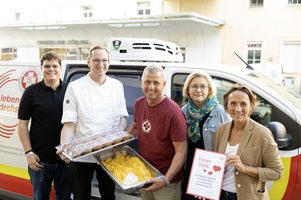 Fünf Personen stehen vor einem PKW des Samariterbund Wien und halten Essen sowie eine Urkunde in der Hand.