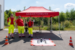 drei Mitarbeiter:innen Samariterbund Wien in Uniform vor einem Scherenzelt, davor Landeplatz einer Drohne 