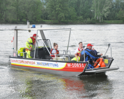 Boot mit Branding von Samariterbund Wien auf Donau, vier Personen in Boot, ein Mann auf linker Seite trägt Samariterbund Uniform und steuert Boot, zwei Frauen mit Schwimmwesten sitzen im Boot und ein Mann in blauem Arbeitsanzug und mit Kappe auf der rechten Seite des Bootes