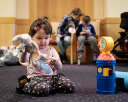 Kleinkind aus Ukraine in Spielraum vom Hotel Ananas, sitzt auf Boden und hält Stofftier in der Hand, daneben ein weiteres Spielzeug, im Hintergrund zwei Burschen, die auf einem Sessel sind und mit dem Handy spielen