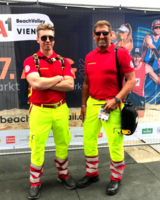 zwei Sanitäter in gelber Uniformhose und rotem Polo-Shirt, beide tragen Brille, lächeln in Kamera, stehen vor Plakatwand Beachvolleyball WM